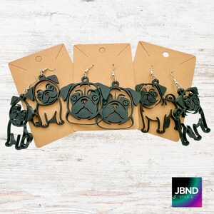 May include: Black pug dog earrings in various poses, displayed on brown cardstock. The earrings are cut from a dark material and feature detailed pug faces and full-body silhouettes. The earrings hang from silver-colored hooks.