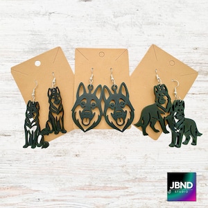 May include: Black German Shepherd dog earrings on display. The earrings are laser cut and feature different poses of the dog breed. The earrings are attached to brown cardstock with silver-colored ear wires. The earrings are displayed on a light wood grain surface.