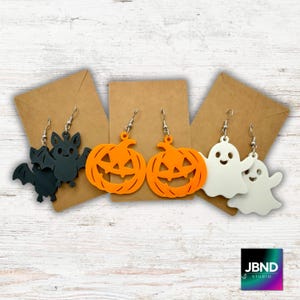 May include: Halloween-themed earrings in the image. There are three pairs of earrings: a black bat, two orange jack-o'-lanterns, and two white ghosts. Each pair is displayed on a brown card against a white wooden background. The earrings have silver hooks.