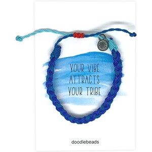 May include: A blue braided bracelet with a small silver charm and a light blue and red accent. The bracelet is displayed on a white card with the text "YOUR VIBE ATTRACTS YOUR TRIBE" and the brand name "doodlebeads".