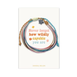 May include: A multi-colored string bracelet with a small flower charm. The bracelet is on a white card with the text "Never forget how wildly capable you are" and "Doodle Beads".