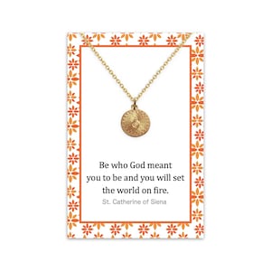Puede incluir: Collar de oro con un colgante redondo con un diseño de sol. El collar viene en una tarjeta con la cita "Sé quien Dios quiso que fueras y encenderás el mundo." de Santa Catalina de Siena.