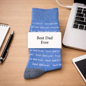 May include: Blue socks with the words "Best Dad Ever" on a white band. The socks are light blue with the words "best dad ever" printed repeatedly. The socks are on a wooden desk next to a laptop, pen, notepad, and smartphone.