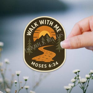 May include: An oval sticker with a nature scene, featuring a sunset over mountains and a winding path. The text "WALK WITH ME" arches across the top, with "MOSES 6:34" at the bottom. The sticker is held by a hand.