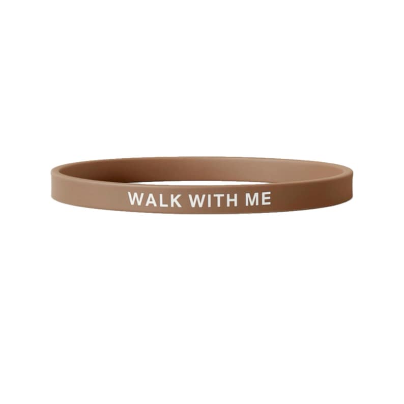 May include: A tan silicone wristband with the words "WALK WITH ME" printed in white. The band is a solid color and has a simple, minimalist design. This item is suitable for everyday wear.