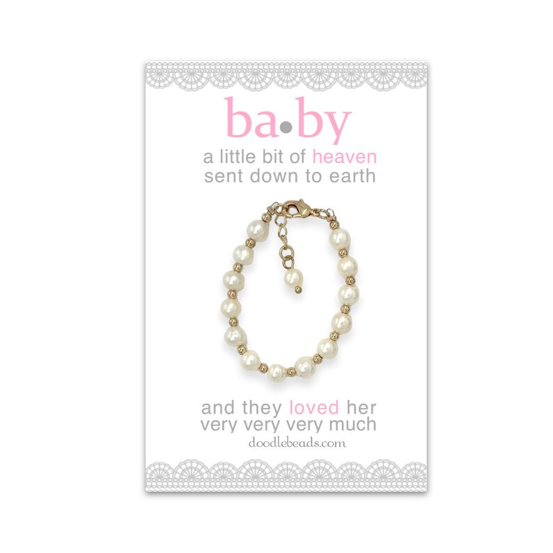 May include: A white pearl bracelet with a gold clasp and a small gold charm. The bracelet is on a white card with the text "ba.by a little bit of heaven sent down to earth and they loved her very very very much doodlebeads.com"