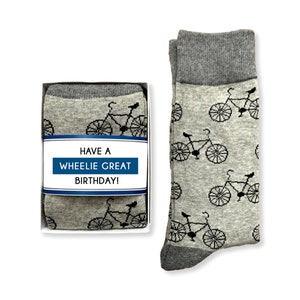 May include: A gray gift box with a pair of gray socks with a black bicycle pattern. The box has a white label with the text "Have a Wheelie Great Birthday!"