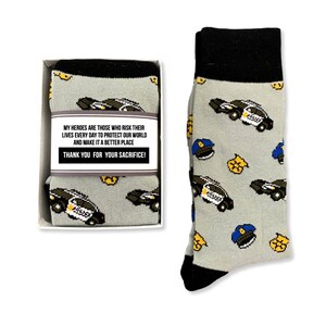 Puede incluir: Calcetines grises con pu&ntilde;o y puntera negros, con un estampado de coches de polic&iacute;a, insignias y gorras. Los calcetines se presentan en una caja blanca con el texto: "MIS H&Eacute;ROES SON AQUELLOS QUE ARRIESGAN SUS VIDAS CADA D&Iacute;A PARA PROTEGER NUESTRO MUNDO Y HACERLO UN LUGAR MEJOR &iexcl;GRACIAS POR SU SACRIFICIO!"