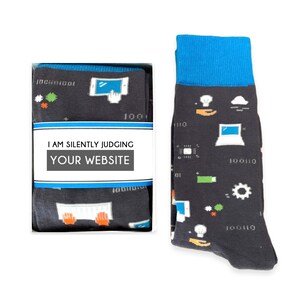 May include: A pair of black socks with a blue cuff. The socks feature a pattern of computer icons, including a laptop, a cloud, a lightbulb, and a gear. The socks are packaged in a white box with the text "I am silently judging your website".