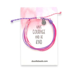 May include: A pink, white, and purple friendship bracelet with a silver charm that says "Have Courage and Be Kind". The bracelet is on a white card with pink watercolor background and the text "doodlebeads.com".