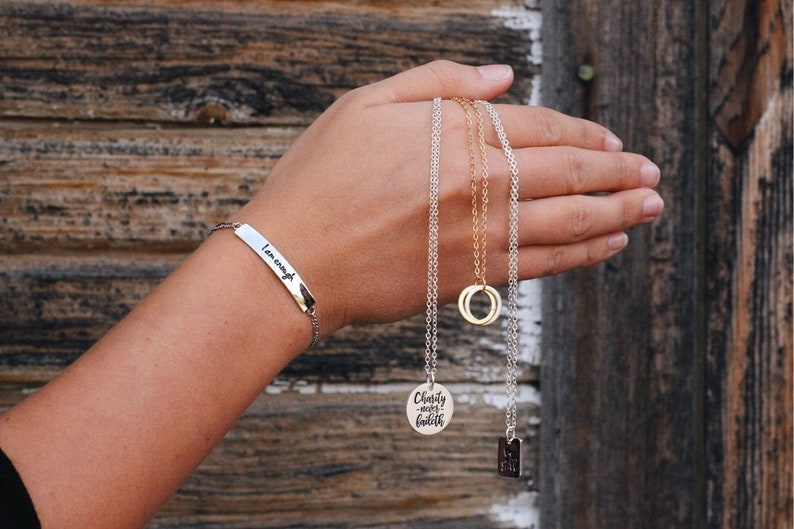 May include: A hand holding three silver necklaces with different pendants. One pendant is a circle, another says "Charity, love, faith", and the third is a small square. The hand is also wearing a silver bracelet that says "I am enough".