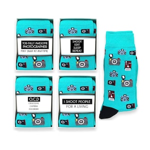 Op de afbeelding: Vier paar turquoise blauwe sokken met een witte rand en een zwarte hiel en teen. Elk paar heeft een ander ontwerp met een camera-thema en tekst. De ontwerpen omvatten "Toe-tally Awesome Photographer May Snap at Anytime", "Shoot Edit Sleep Repeat", "OCD Obsessive Camera Disorder", en "I Shoot People For A Living".