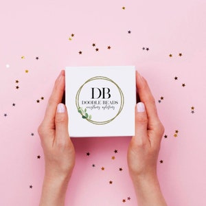 May include: A white box with a gold circle logo that says "DB Doodle Beads everything uplifting" on a pink background with gold stars.