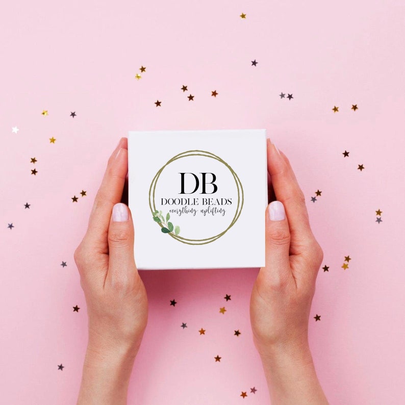 May include: A white box with a gold circle logo that says "DB Doodle Beads everything uplifting" on a pink background with gold stars.
