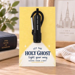 May include: A black cylindrical flashlight with a carabiner clip attached to a yellow display card. The flashlight features the text "SHARE YOUR LIGHT". The card displays the text "LET THE HOLY GHOST light your way SHARE YOUR LIGHT" with star icons.