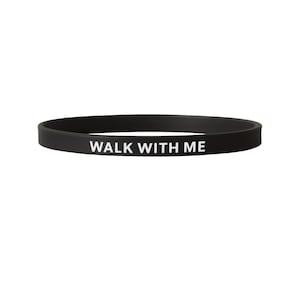May include: A black silicone wristband with the white text "WALK WITH ME". The band is a simple, circular design, suitable for everyday wear. The text is centered and clearly visible against the dark background.