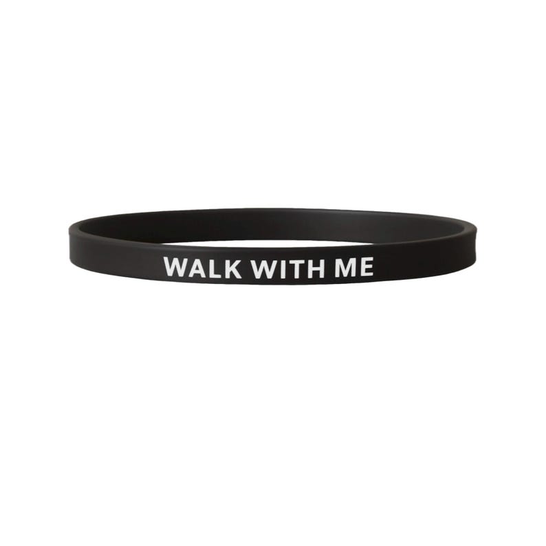 May include: A black silicone wristband with the white text "WALK WITH ME". The band is a simple, circular design, suitable for everyday wear. The text is centered and clearly visible against the dark background.