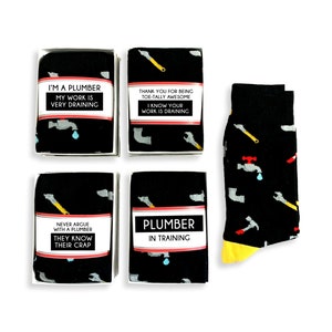 Puede incluir: Cuatro cajas de regalo negras con bordes blancos contienen calcetines con temática de fontanero. Los calcetines tienen un fondo negro con herramientas de fontanería blancas, amarillas y rojas y una gota de agua azul. Los calcetines están empaquetados en cajas con etiquetas blancas que tienen dichos divertidos sobre los fontaneros. Los dichos son: "Soy fontanero, mi trabajo es muy agotador", "Gracias por ser tan genial, sé que tu trabajo es agotador", "Nunca discutas con un fontanero, ellos saben su trabajo", y "Fontanero en formación".