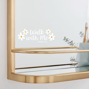 May include: A gold-framed mirror with a shelf. The mirror has the text "Walk with Me" and the reference "Moses 6:34" in white, along with daisy graphics. The shelf is holding a white container and a toothbrush.