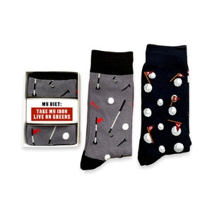 May include: Two pairs of socks with a golf theme. One pair is gray with white golf balls, golf clubs, and red flags. The other pair is navy blue with white golf balls and red flags. The socks are packaged in a white box with the text "My Diet: Take My Iron Live On Greens".