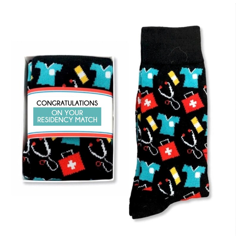 May include: A gift box containing a pair of black socks with a pattern of medical symbols, including a stethoscope, a first aid kit, and a doctor's coat. The box has a white label with the text "Congratulations on your residency match".