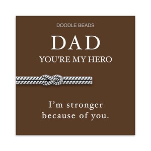 May include: A brown card with the text "DAD YOU'RE MY HERO" and "I'm stronger because of you." A silver-coloured knot-shaped accessory is featured below the text. The brand name "DOODLE BEADS" is at the top.