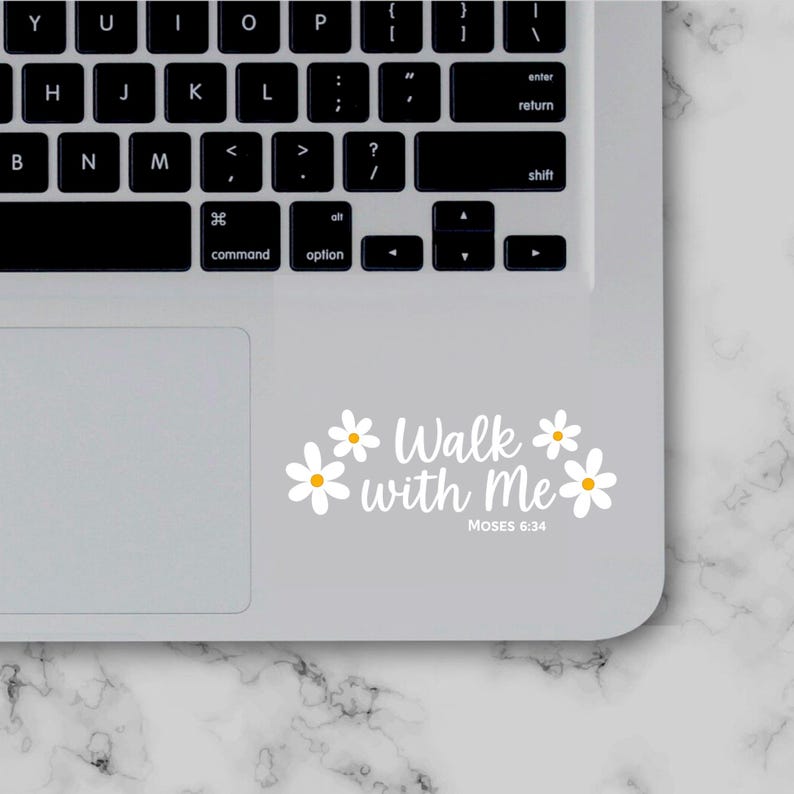 May include: A white sticker with the text "Walk with Me" and the reference "Moses 6:34" in a script font, surrounded by white daisy illustrations. The sticker is placed on a laptop.