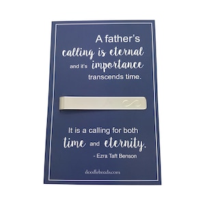May include: A silver tie bar with an infinity symbol engraved on it. The tie bar is on a blue card with the text "A father's calling is eternal and it's importance transcends time. It is a calling for both time and eternity. - Ezra Taft Benson doodlebeads.com"