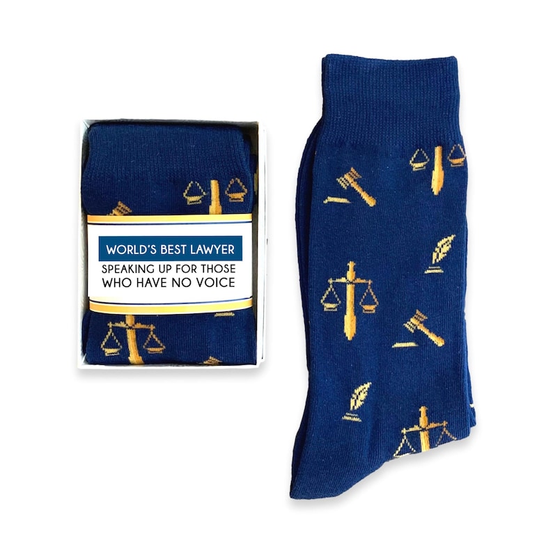 May include: A gift box containing a pair of blue socks with a gold scale of justice, gavel, and quill pattern. The box has a white label that reads "World's Best Lawyer Speaking Up For Those Who Have No Voice."