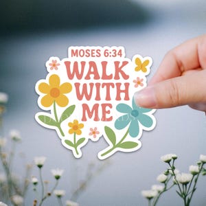May include: A sticker with the text "MOSES 6:34 WALK WITH ME" in a retro font, surrounded by colorful flowers and a butterfly. The sticker is held by a hand against a blurred background.