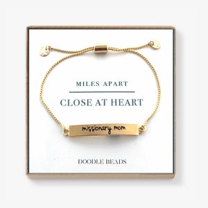 May include: Gold adjustable bracelet with a small gold bar charm that reads "missionary mom". The bracelet is in a box with the words "Miles Apart Close At Heart" and "Doodle Beads" printed on it.