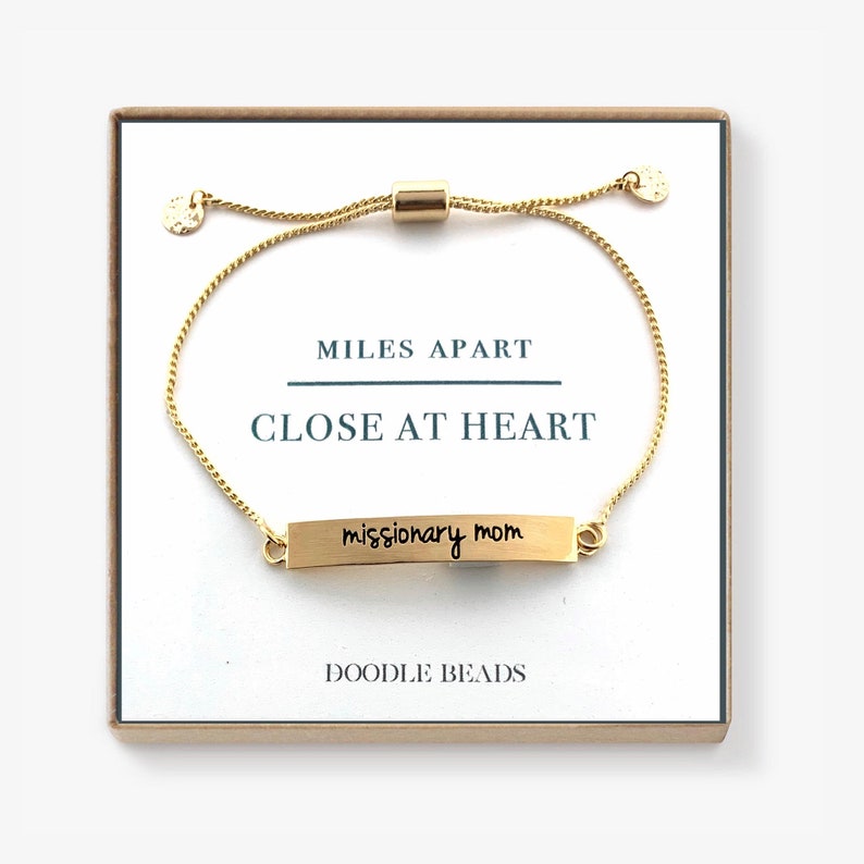 May include: Gold adjustable bracelet with a small gold bar charm that reads "missionary mom". The bracelet is in a box with the words "Miles Apart Close At Heart" and "Doodle Beads" printed on it.