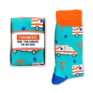 May include: A pair of blue socks with a pattern of white ambulances and red medical symbols. The socks are packaged in a white box with the text "Paramedic More Than Band-Aids For Boo-Boos".