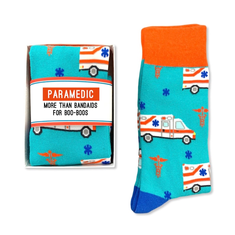 May include: A pair of blue socks with a pattern of white ambulances and red medical symbols. The socks are packaged in a white box with the text "Paramedic More Than Band-Aids For Boo-Boos".