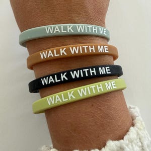 May include: A collection of four silicone wristbands in various colors: sage green, brown, black, and lime green. Each band features the white text "WALK WITH ME". The bands are stacked on a wrist, showcasing the different color options.