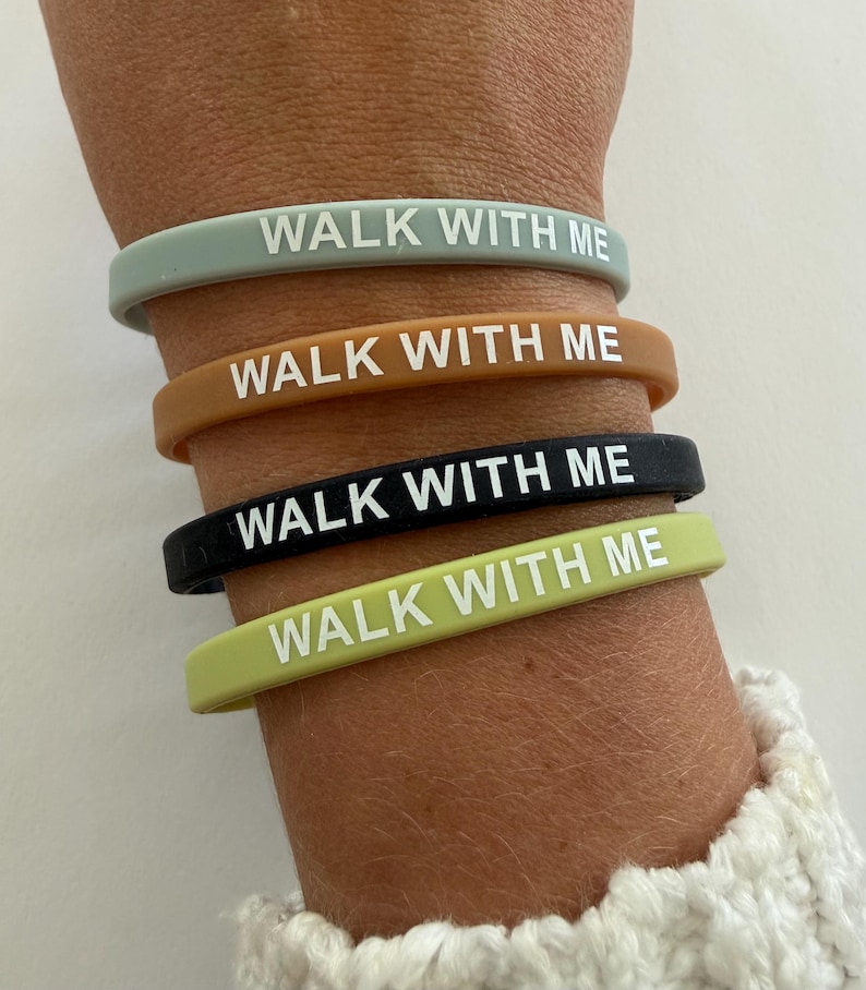 May include: A collection of four silicone wristbands in various colors: sage green, brown, black, and lime green. Each band features the white text "WALK WITH ME". The bands are stacked on a wrist, showcasing the different color options.
