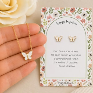 May include: A gold-colored butterfly necklace and matching earrings with white accents are displayed next to a card that says "happy baptism." The card has a floral design and a quote about baptism.