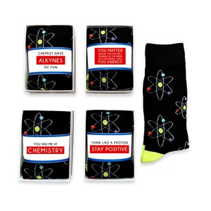 Peut inclure: Quatre petites boîtes cadeaux avec des motifs d'atomes noirs et blancs et des dictons amusants sur le thème des sciences. Les boîtes sont disposées en deux rangées de deux. Les dictons sont : "Chemist Have Alkynes of Fun", "You Matter Unless You Multiply Yourself by the Speed of Lightening Then You Energy", "You Had Me at Chemistry", et "Think Like a Proton Stay Positive". Une chaussette noire avec un motif d'atome blanc est également représentée.