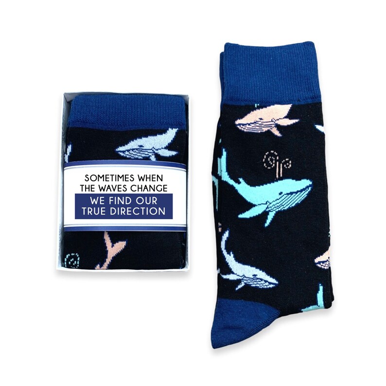 May include: A pair of black socks with a blue cuff and toe, featuring a pattern of light blue and pink whales. The socks are displayed next to a box with the text "SOMETIMES WHEN THE WAVES CHANGE WE FIND OUR TRUE DIRECTION."