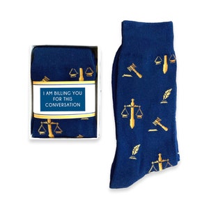 May include: A pair of blue socks with a gold scale of justice, gavel, and quill pattern. The socks come in a white box with a label that reads "I am billing you for this conversation."