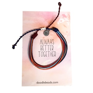 Könnte beinhalten: Buntes Freundschaftsarmband mit einem silbernen Herz-Anhänger. Das Armband wird auf einer Karte mit den Worten "Always Better Together" gezeigt.