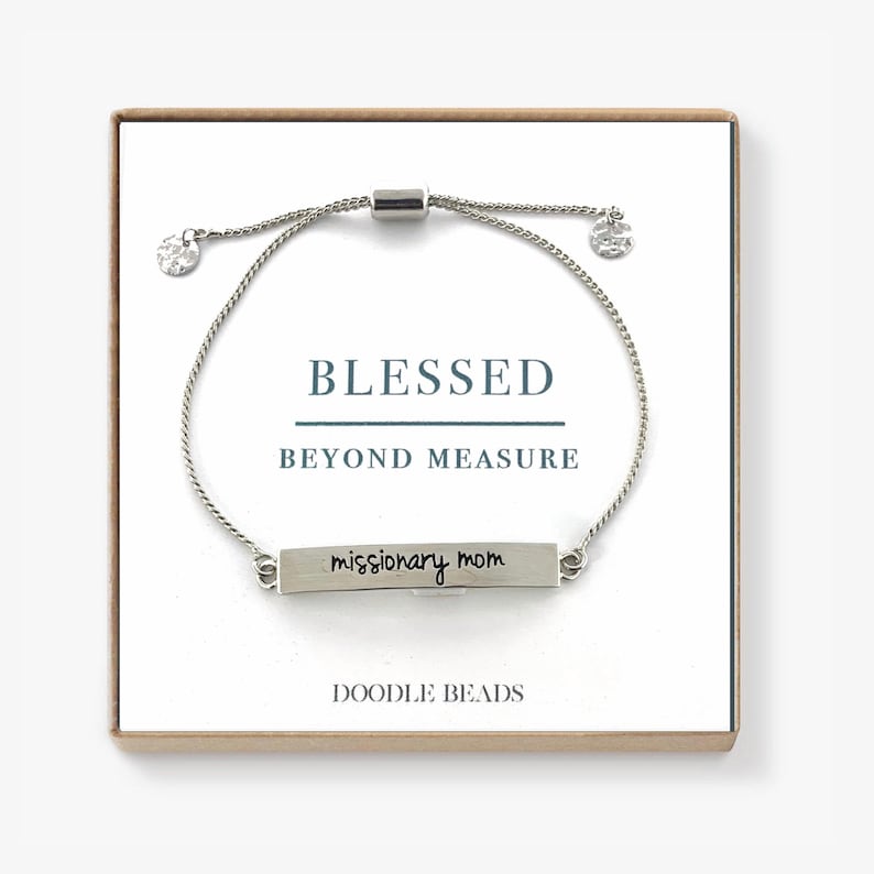May include: Silver bracelet with a silver bar charm that says "missionary mom" and a silver slider bead. The bracelet is in a box with the words "Blessed Beyond Measure" and "Doodle Beads" printed on it.