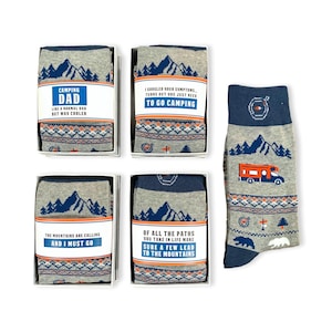 以下が含まれることがあります： 青いグレーの靴下が入った白い箱が4つ。靴下は山とキャンプをテーマにしている。箱には「Camping Dad」、「To Go Camping」、「The Mountains Are Calling And I Must Go」、「Of All The Paths You Take In Life Make Sure A Few Lead To The Mountains」という文字が書かれている。