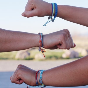 May include: Three wrists with colorful braided bracelets. The bracelets are made of different colored threads and have silver charms.