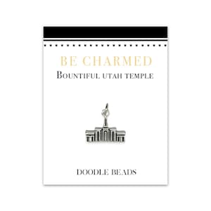 Puede incluir: Un amuleto de plata que representa una representación detallada del Templo de Bountiful, Utah. El amuleto se muestra en una tarjeta blanca con el texto "BE CHARMED" y "BOUNTIFUL UTAH TEMPLE" en letras negras. La tarjeta también incluye el texto "DOODLE BEADS" en letras negras.