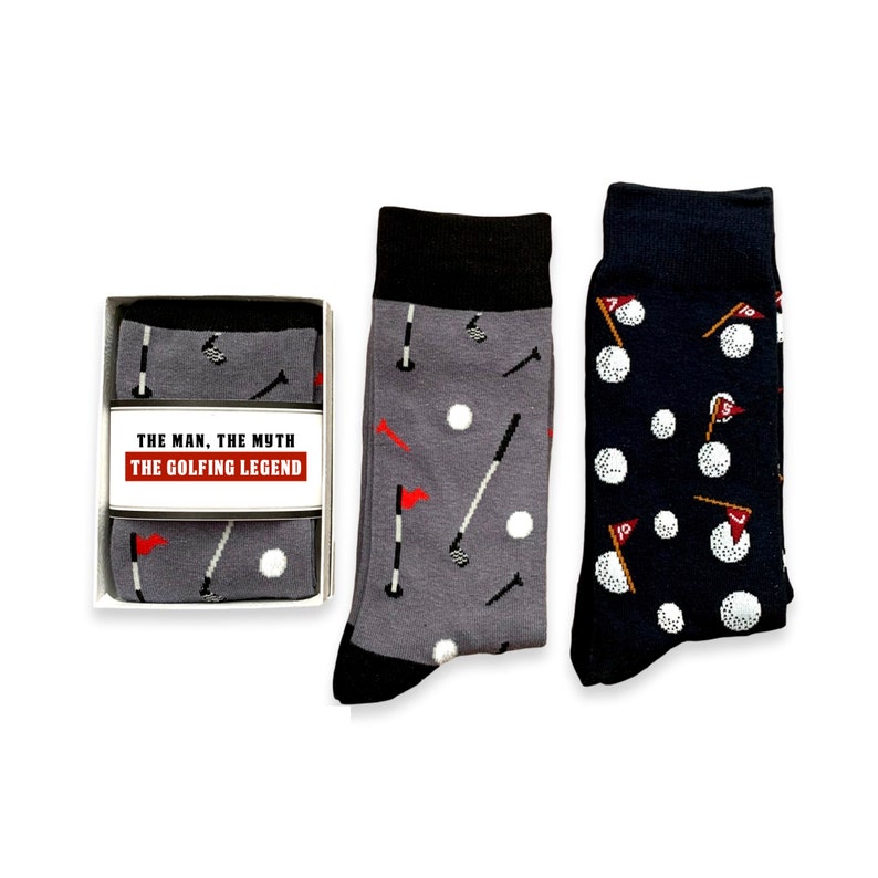 May include: Two pairs of socks with a golf theme. One pair is gray with white golf balls, golf clubs, and red flags. The other pair is navy blue with white golf balls and red flags. The socks are packaged in a white box with the text "The Man, The Myth, The Golfing Legend".