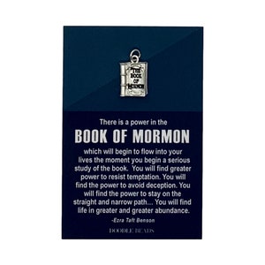 May include: A silver charm in the shape of a book with the words "The Book of Mormon" on the cover. The charm is on a card with a quote from Ezra Taft Benson about the power of the Book of Mormon. The card is blue with white text and the words "Doodle Beads" at the bottom.
