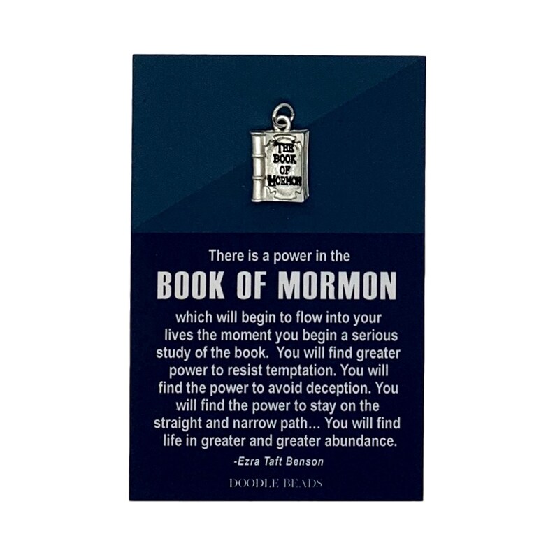 May include: A silver charm in the shape of a book with the words "The Book of Mormon" on the cover. The charm is on a card with a quote from Ezra Taft Benson about the power of the Book of Mormon. The card is blue with white text and the words "Doodle Beads" at the bottom.