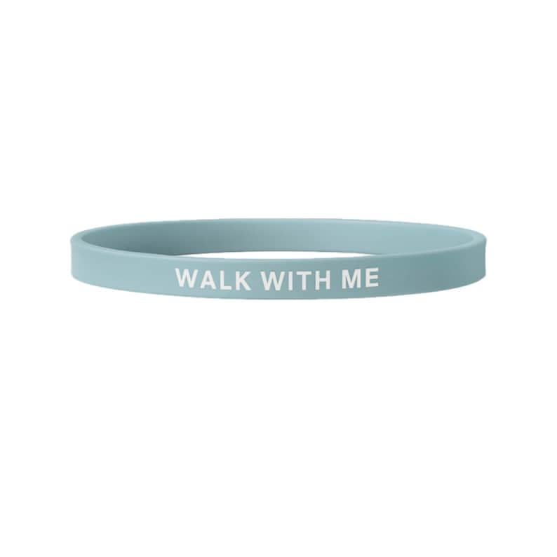 May include: A light blue silicone wristband with the phrase "WALK WITH ME" printed in white. The band is a simple, circular design, suitable for everyday wear. The wristband is approximately 8 inches in circumference.