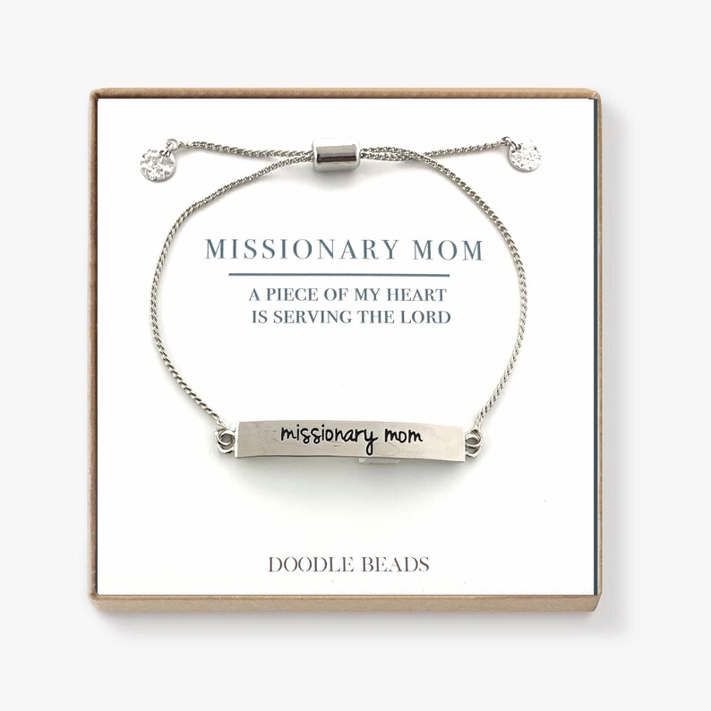 May include: Silver bracelet with a silver bar charm that says "missionary mom" in cursive. The bracelet is on a white background with a card that says "Missionary Mom, A piece of my heart is serving the Lord, Doodle Beads".